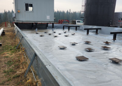 Industrial containment area with a geomembrane liner, support brackets, and mounted platforms, surrounding a vertical storage tank and elevated control building in a remote worksite.